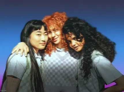Three young, diverse women standing close together with their arms around each other against a blue background with a pink gradient at the bottom. They are facing the camera with relaxed expressions and are wearing light-coloured tops layered with patterned sweater vests.