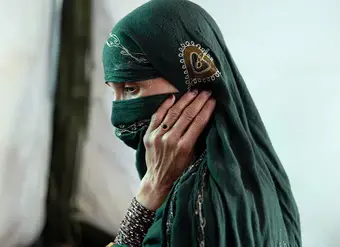 Close-up portrait of a woman in a dark green veil, with her hand resting against her face.