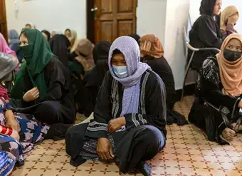 Women sit together on the floor inside an indoor space, with a woman wearing a purple headscarf seated in the foreground.