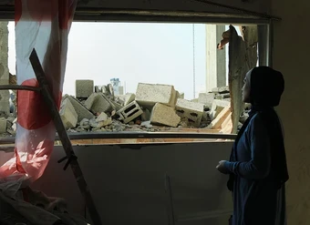 A woman looks out at rubble from inside a ruined building.