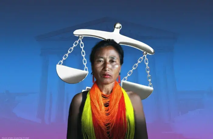 Portrait of a woman standing against a blue-to-purple gradient background with a faint image of a classical courthouse-style building with columns behind her. A large silver balance scale appears above and behind her head, with one side tilted lower than the other. She is wearing a black top and layered beaded necklaces in bright orange, yellow and red. She faces the camera with a serious expression.