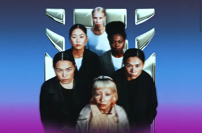 Six women standing closely together against a blue-to-purple gradient background, facing the camera with serious expressions. They are positioned in two rows, with a metallic UN Women logo displayed behind them. The women have diverse appearances and wear dark and neutral-toned clothing, creating a strong, unified group portrait.