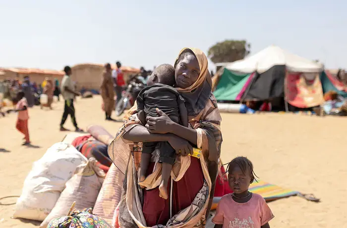 A woman holds a child and looks at the camera, with belongings behind her and another child beside her.