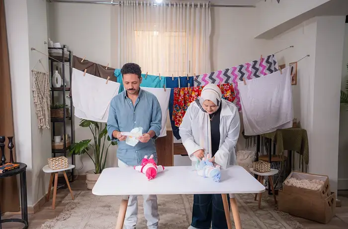 Dos adultos están de pie junto a una mesa cambiando los pañales a unas muñecas, con la ropa tendida en un tendedero detrás de ellos, en un entorno doméstico.