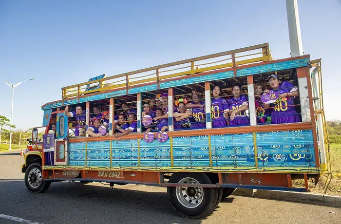 Un grupo de mujeres con camisetas moradas viajan juntas en una furgoneta abierta por los lados, sonriendo y sosteniendo carteles.