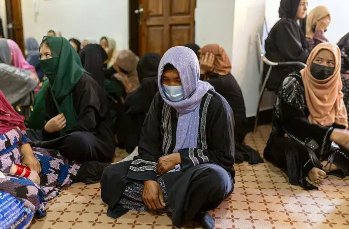 Women sit together on the floor inside an indoor space, with a woman wearing a purple headscarf seated in the foreground.