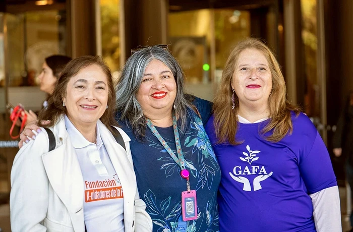 Three women pose together outside a building, with two wearing purple shirts featuring organization logos.
