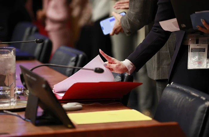 A person at a meeting table holds a document beside microphones and other meeting materials