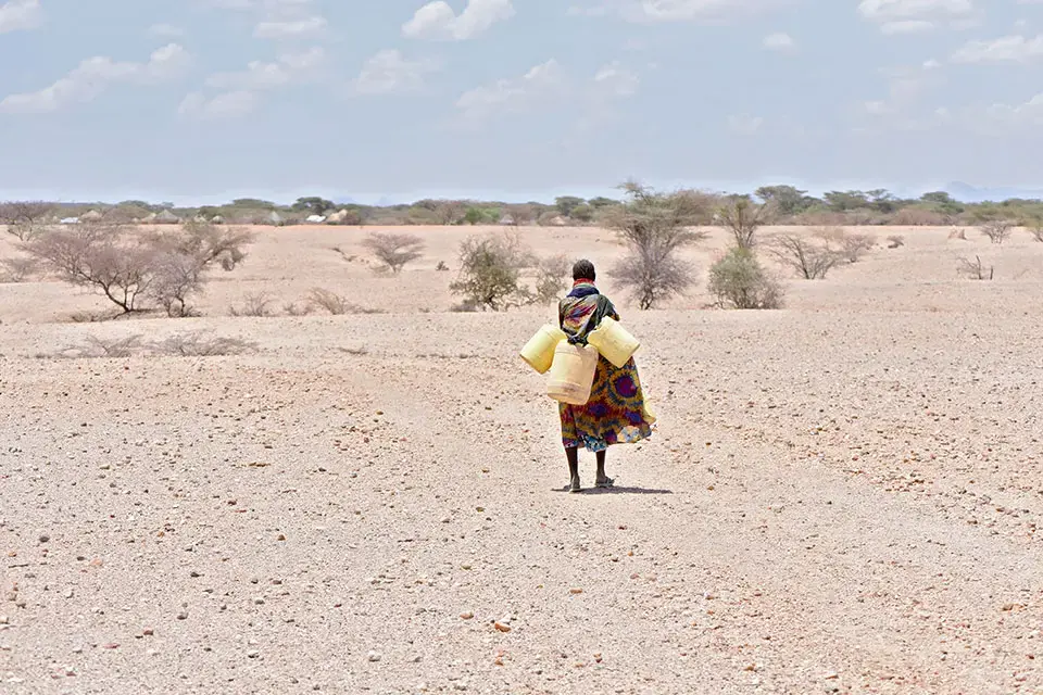 A women carries three large empty water jugs over arid land
