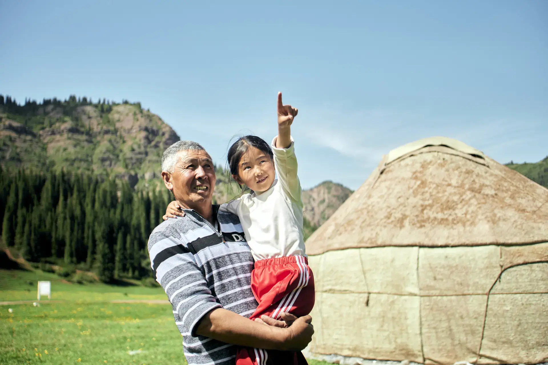 A man carries a girl who points toward the sky while standing in a grassy landscape with mountains and a tent in the background.