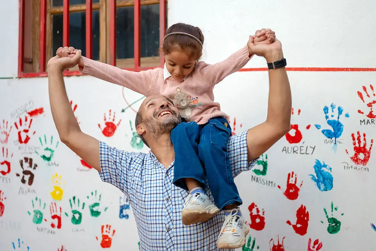 Father carries his daughter onto his shoulders while they play in front of a wall decorated with colourful handprints.