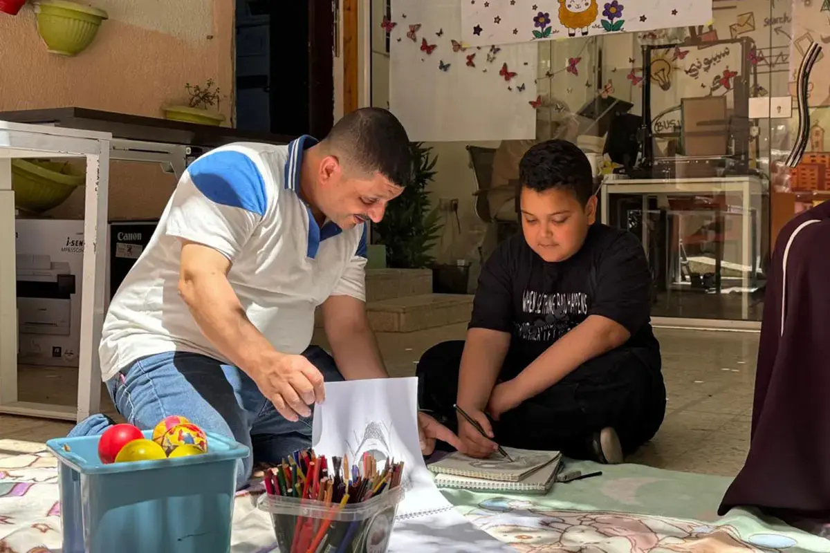 Father and son sit on the floor drawing together, surrounded by art supplies and toys.