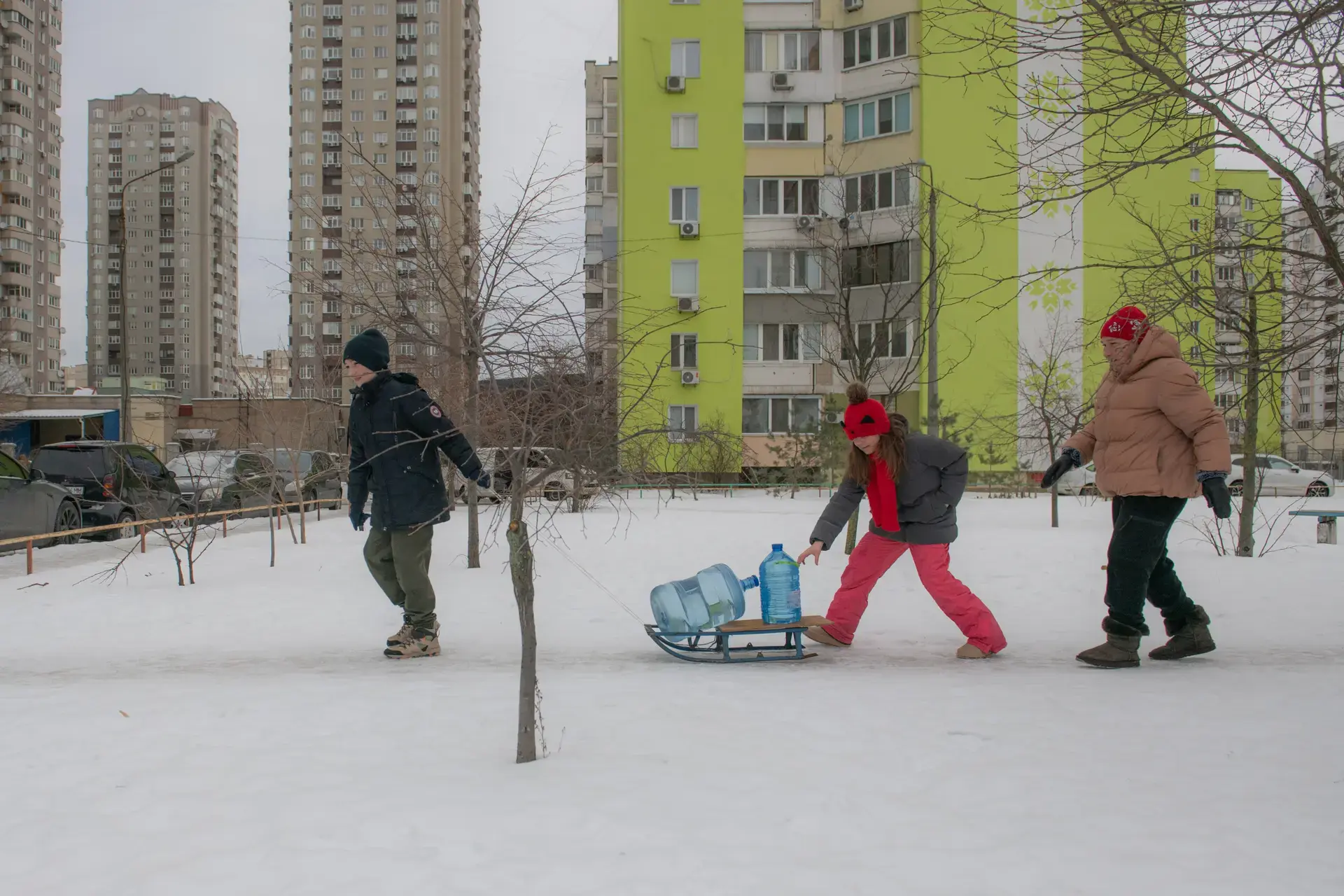 Three people walk through a snowy residential area, pulling large water containers on a sled between apartment buildings.