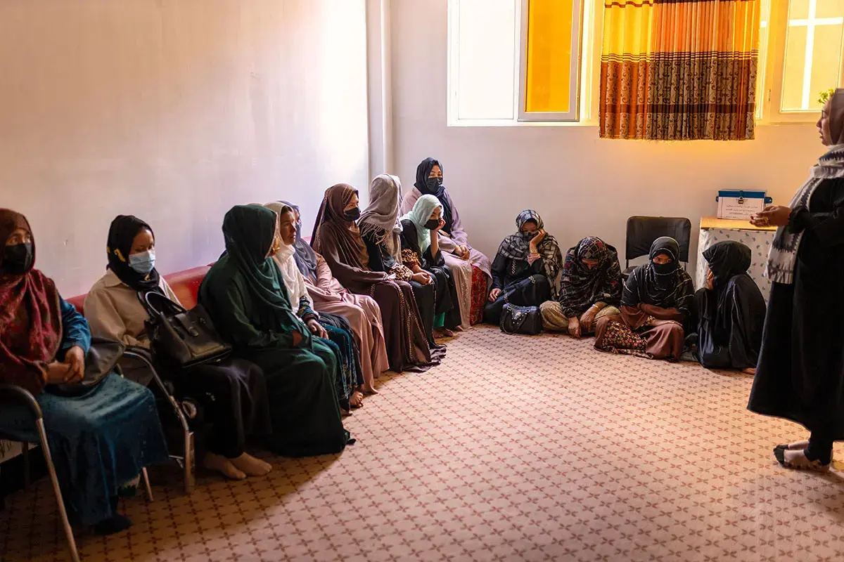 Women sit together inside an indoor space while a facilitator speaks to the group.