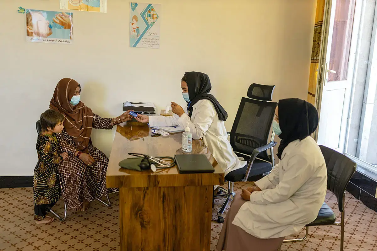 A health worker provides a medical consultation to a woman while a child stands beside her in a clinic room, with another health worker seated nearby.