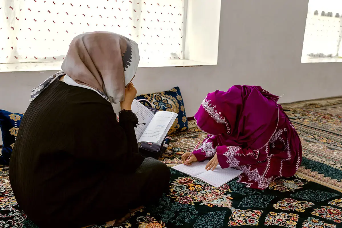 A woman sits beside a girl who is writing in a notebook on the floor of an indoor space.