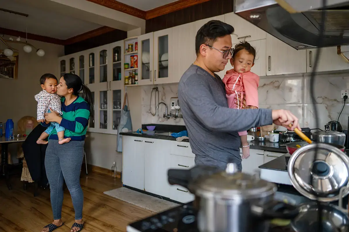 In Nepal, a public information campaign aims to make shared caregiving among partners the norm rather than the exception. When parents share caregiving and household responsibilities, they also become positive role models for their children. In this photo, a man cooks while holding a child, while a woman is attending to another child in the family kitchen.  child, as a woman holds another child in the family kitchen. Photo: UN Women/Srijana Bhatta