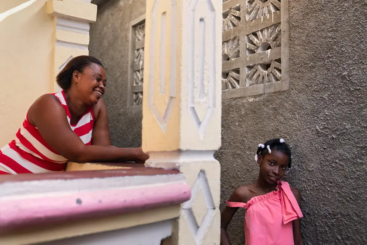 A smiling woman leans on a porch railing beside a girl in a pink dress.
