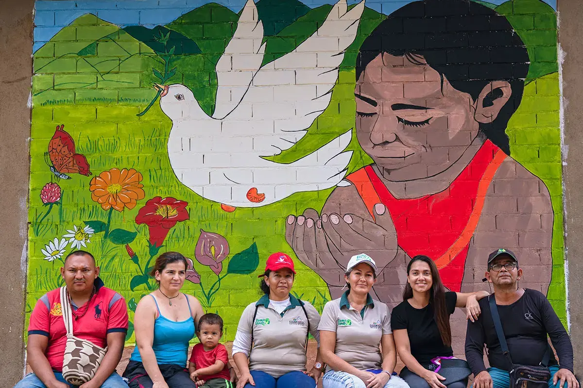 Grupo de cuatro mujeres, dos hombres y un niño sentados frente a un mural de paz