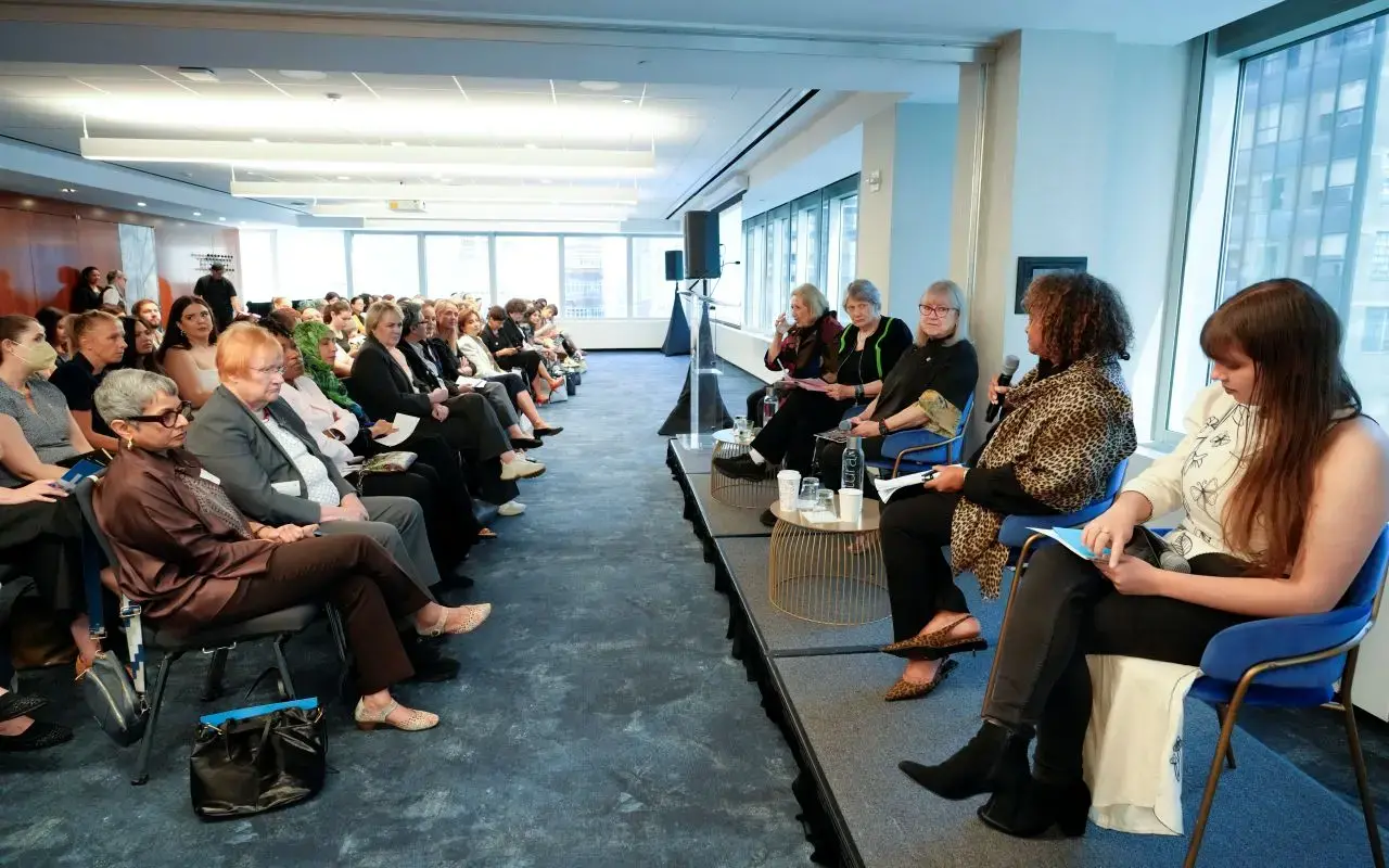 A panel of five women speaks in front of a full audience