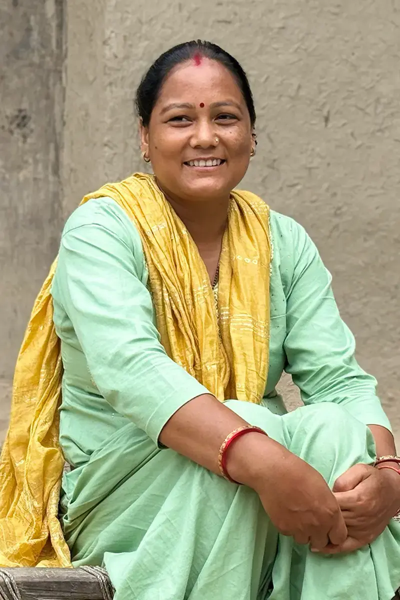 A portrait of a smiling woman wearing a light green outfit and a yellow scarf, seated outdoors against a plain wall.