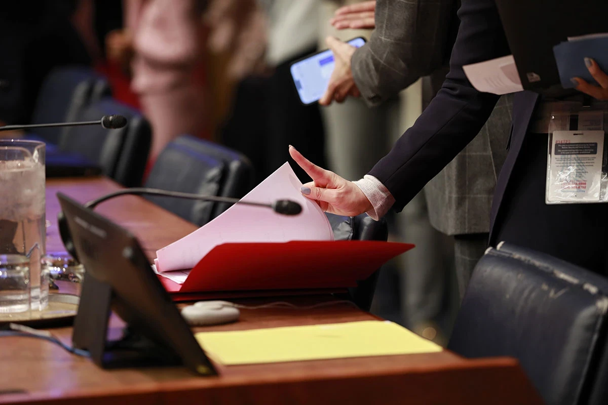 A person at a meeting table holds a document beside microphones and other meeting materials