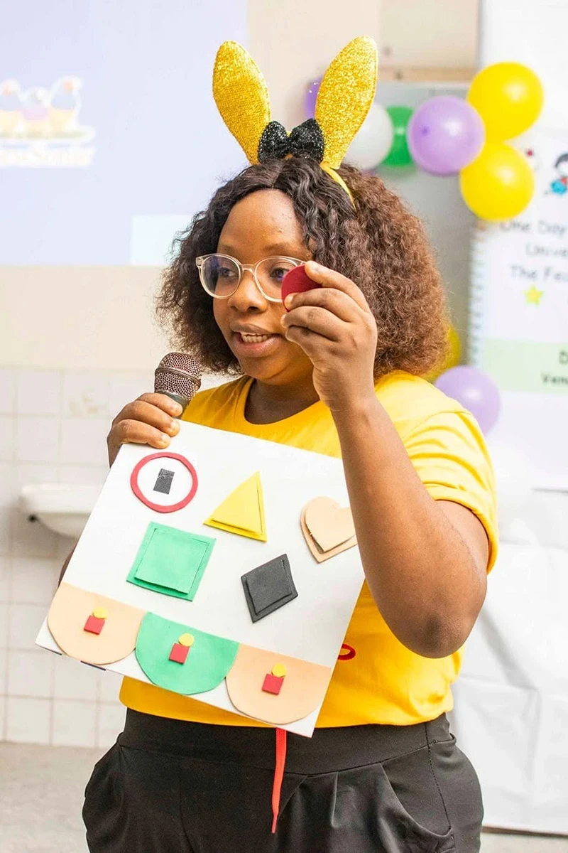 A woman speaks into a microphone while holding a visual aid with coloured shapes, wearing a headband with bunny ears.