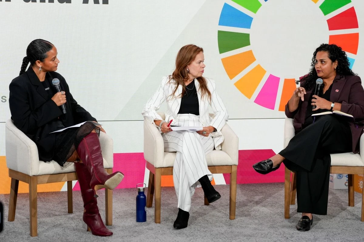 Three women speak on a stage in front of the colorful logo of the SDGs